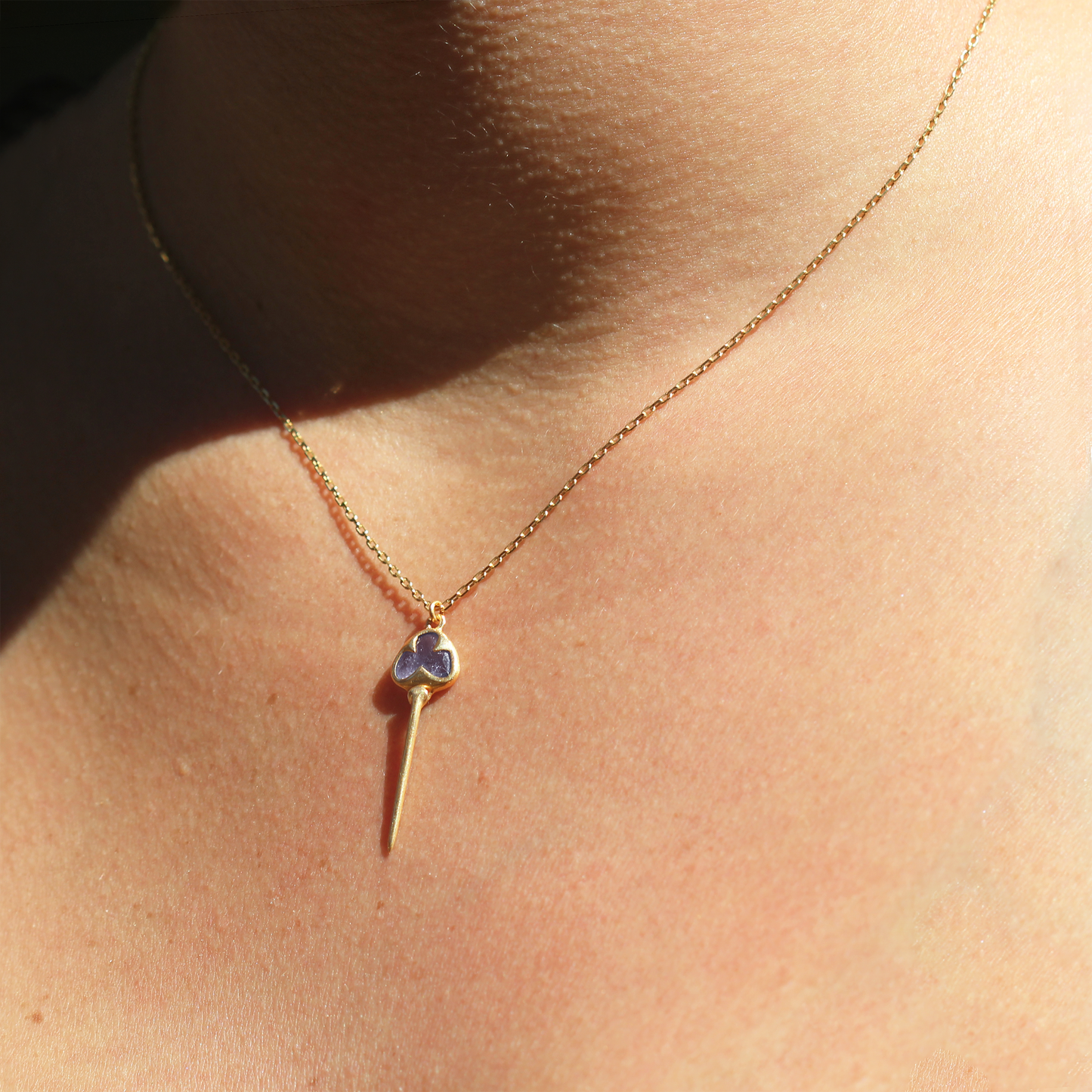 Woman wearing Yellow gold necklace with bezel set, pale purple tanzanite stone, and an elongated gold stingray tail dangling from the bottom, on a dainty gold chain