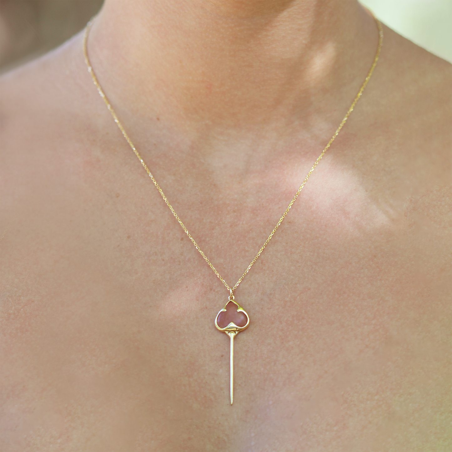 Woman wearing Yellow gold necklace with bezel set muted pink guava quartz stone, and an elongated gold stingray tail dangling from the bottom, on a dainty gold chain