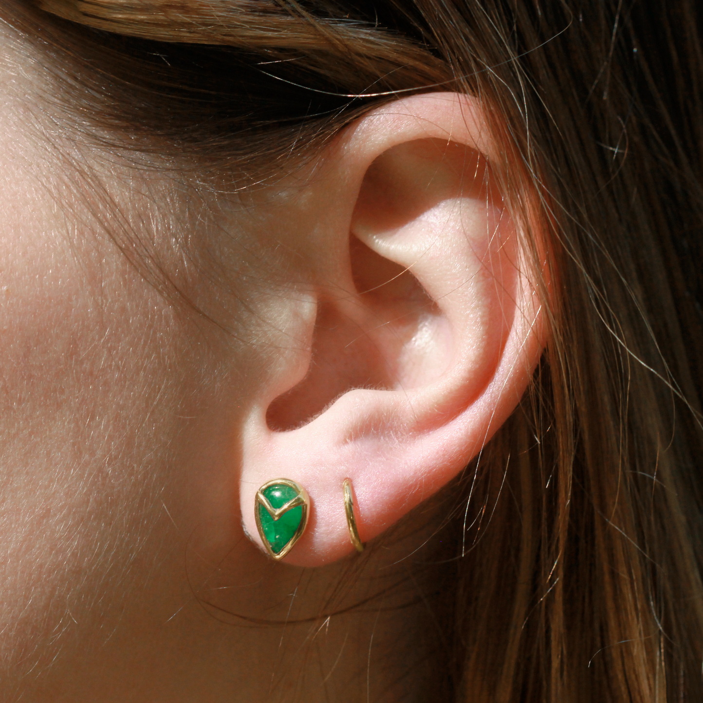 Woman wearing A pair of stud earrings with teardrop-shaped, green emerald stone, bezel set with yellow gold owl beak detail