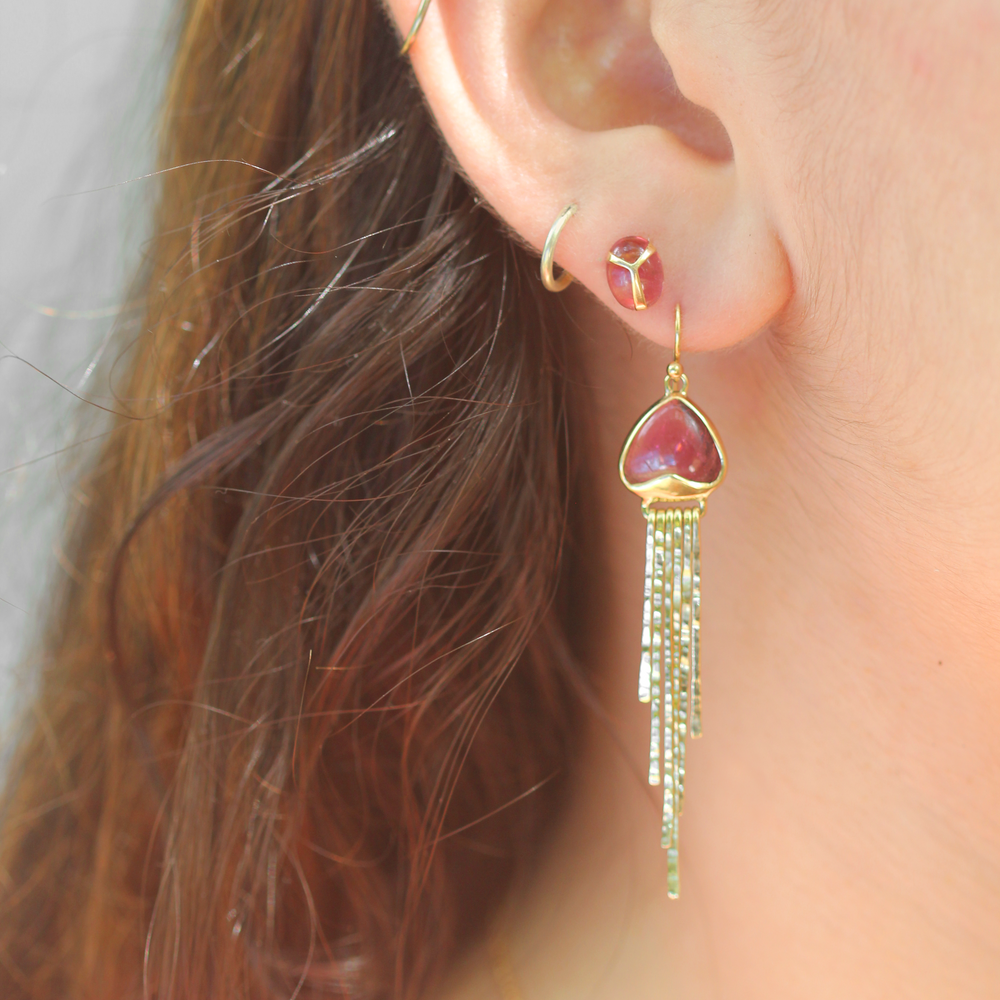 Closeup of woman wearing Dangle earrings in Pink Tourmaline and 18k Yellow Gold. Bezel set, triangle-shaped stone, narrow hammered gold dangling elements, and french hooks.